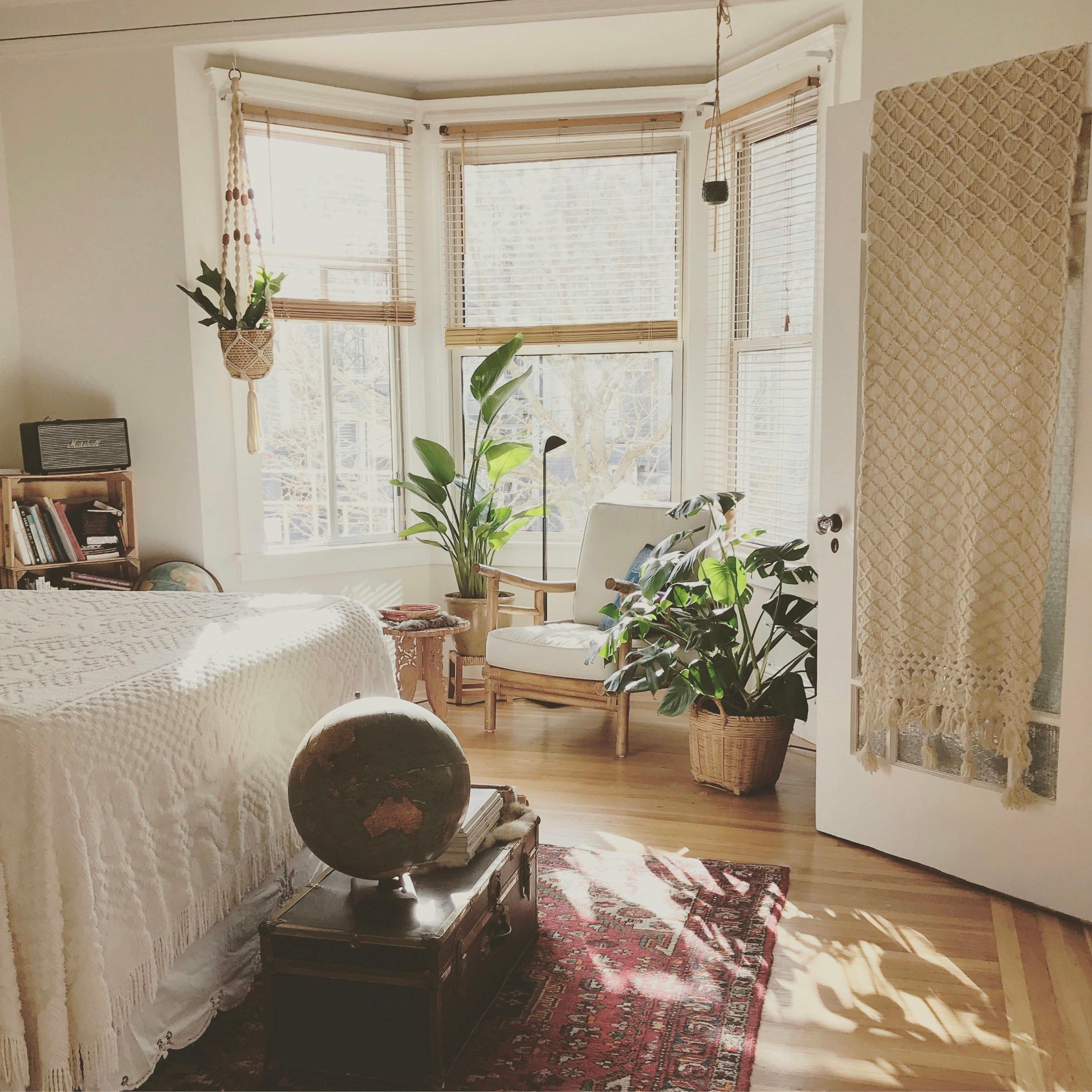 Cozy NYC bay-windowed bedroom with macrame, vintage globe, woven blankets, and a hanging plant in soft natural light