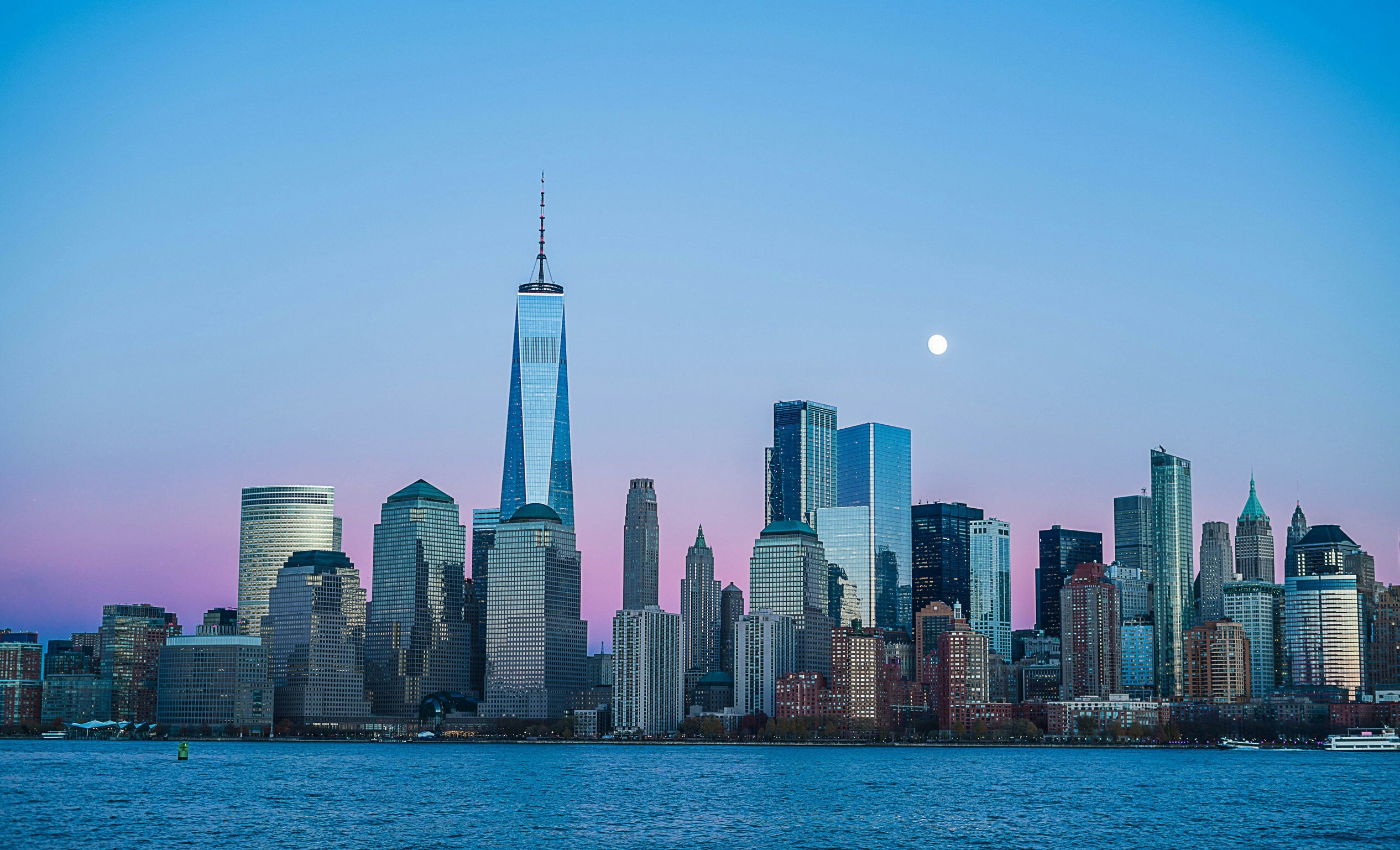 Downtown Manhattan skyline at dusk with One World Trade Center, a rising moon, and pink-purple sky reflected in the harbor